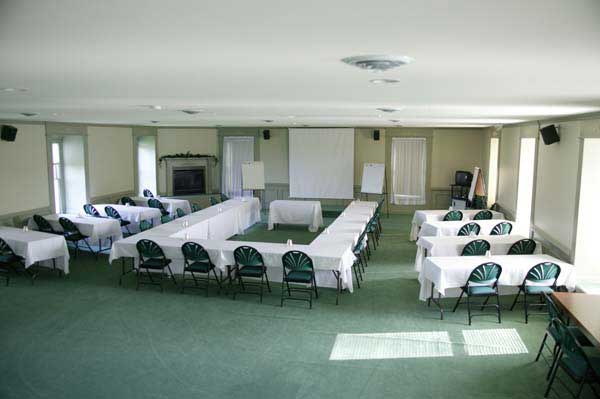 An empty conference room with tables arranged in a U-shape, covered with white tablecloths and surrounded by green cushioned chairs; there is a fireplace on one end and whiteboards on the other, with natural light coming through windows.