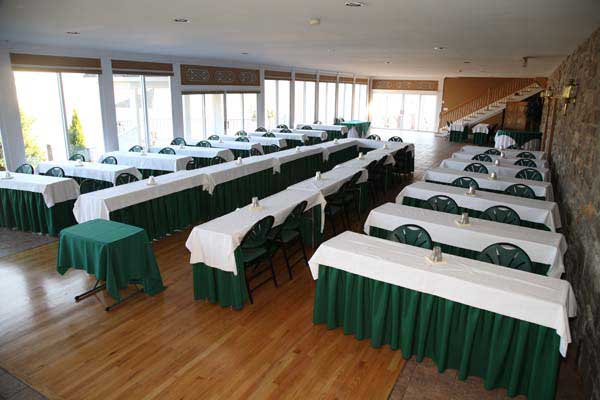 A banquet hall with rows of white-covered tables with green skirting and empty green-cushioned chairs set up for an event.