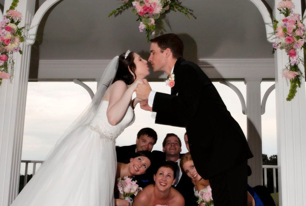 Bride and groom kiss on pavilion with wedding party watching