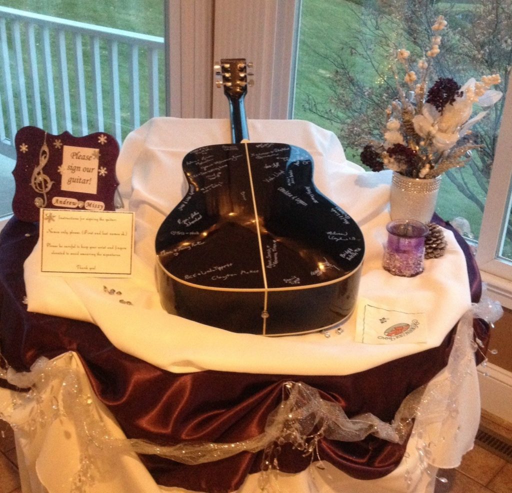 A table covered in white and purple satin holds a black guitar, which is positioned upside down and decorated with signatures. Next to the guitar—a unique wedding guest book idea—is a sign with a musical note design, a small vase with pine cones, and a purple candle in a glass holder.