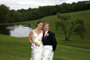 A wedding couple stands on a grassy hill in front of a serene pond. The bride is in a white gown holding a bouquet of white flowers, and the groom is in a dark suit with a red tie. Both are smiling with green trees and rolling hills in the background.