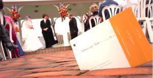 A winter wedding ceremony unfolds outdoors, with a bride and groom standing at the altar. Chairs with guests are seen in the background and foreground, and a wedding program, presumably detailing the event, lies on the brick floor in the foreground.