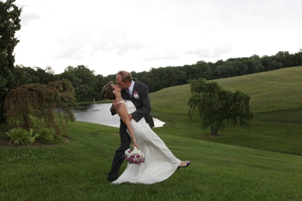 Groom dips the bride as she hold her bouquet of flowers on the back lawn in front of the pond and old willow treel