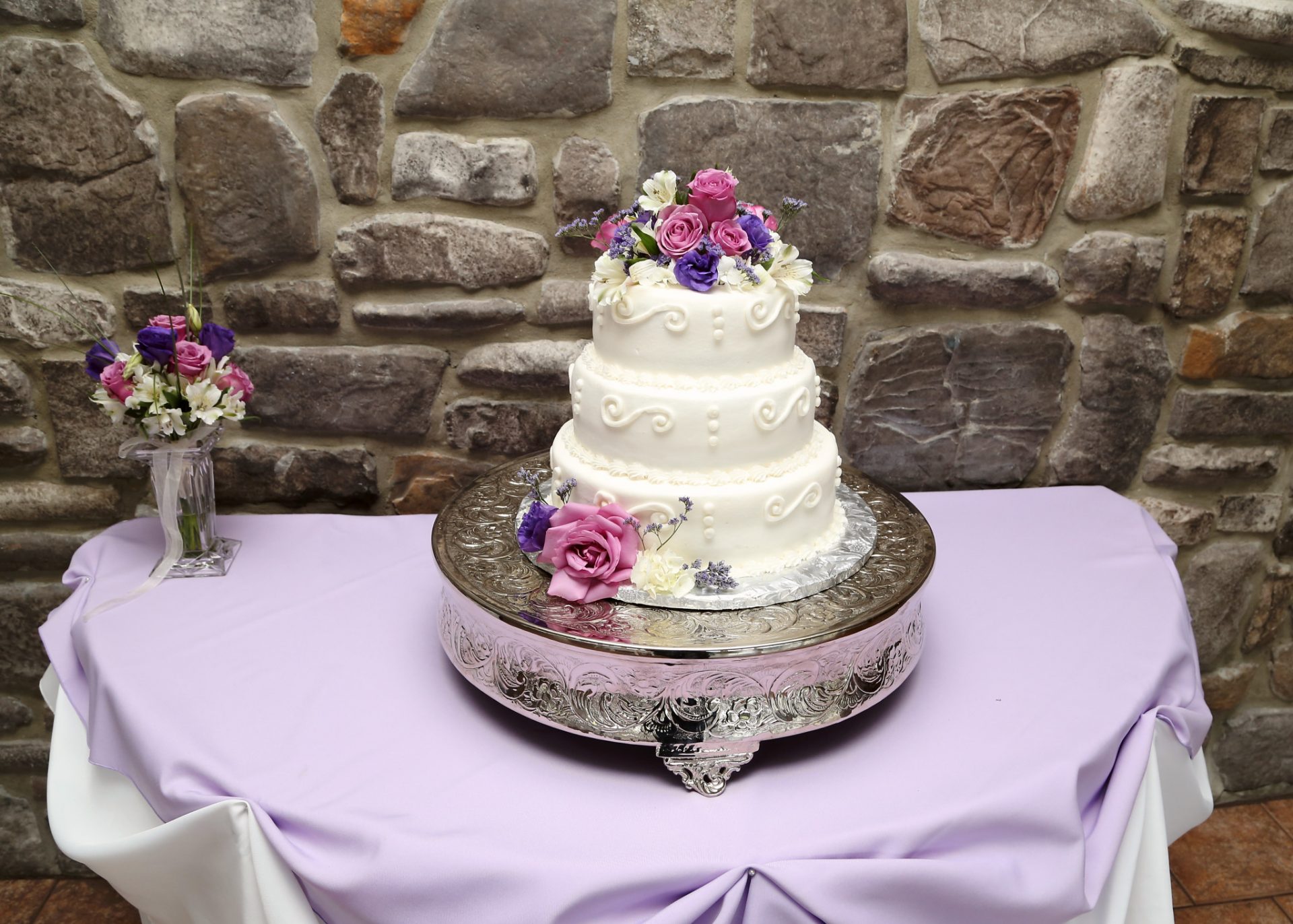Three tiered wedding cake with white cream icing and decorated with pink and purple flowers. The cake sits on a silver cake display stand.