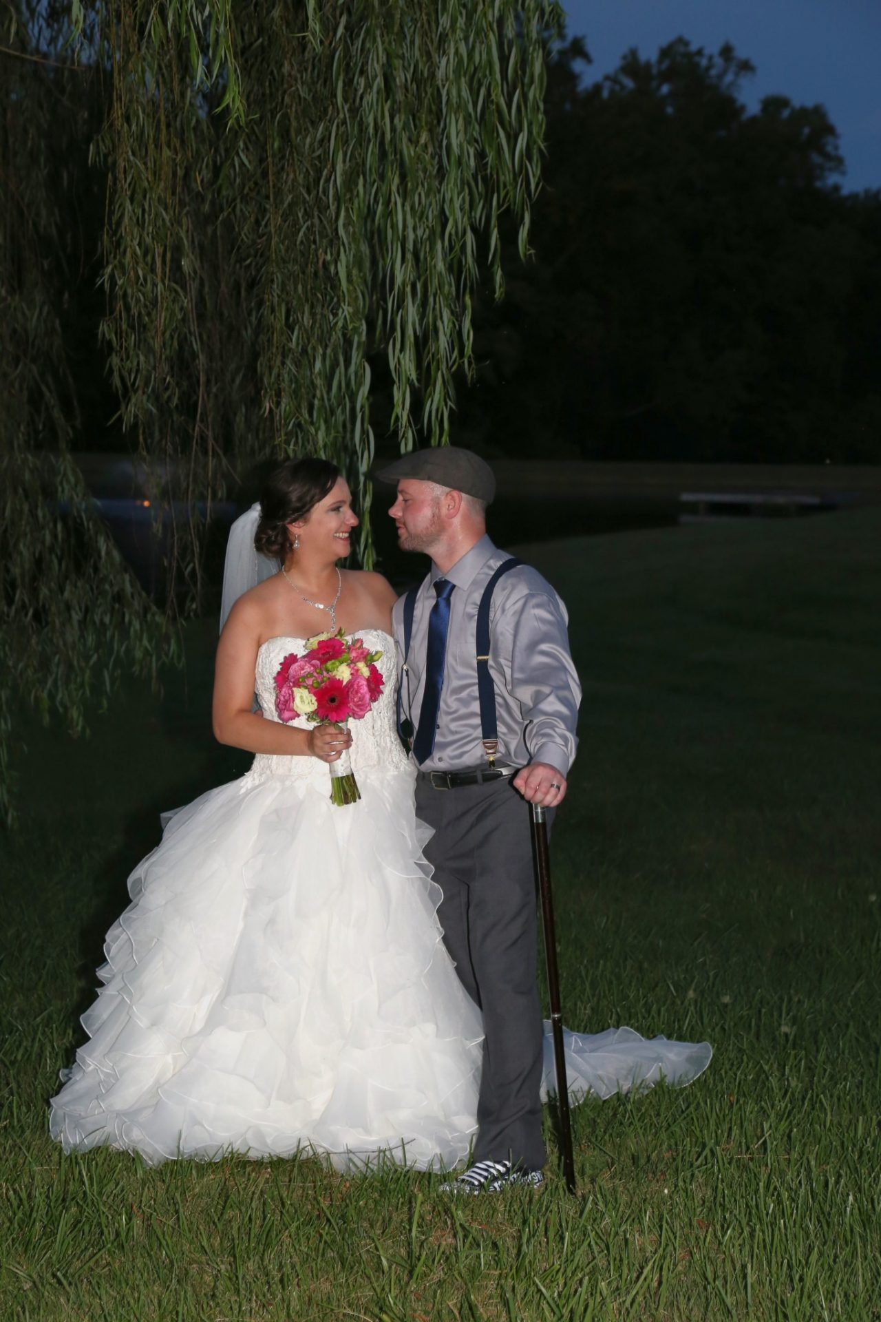 Bride and groom pose by old willow tree by pond.