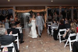 A bride and groom walking down the aisle while guests seated on either side are looking on, some clapping and capturing the moment on their phones. The venue is decorated with white chairs, petals on the ground, and string lights around the windows.