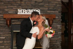 A newlywed couple kissing in front of a stone fireplace with a sign above them reading "MR & MRS." The bride is holding a bouquet of colorful flowers, and both are dressed in traditional wedding attire.