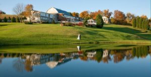 A serene, scenic view featuring a large house on a lush, green hillside with trees in autumn colors. A couple dressed in wedding attire stands near the water which reflects the house, trees, and gazebo near the shore under a clear blue sky. | Morningside Inn