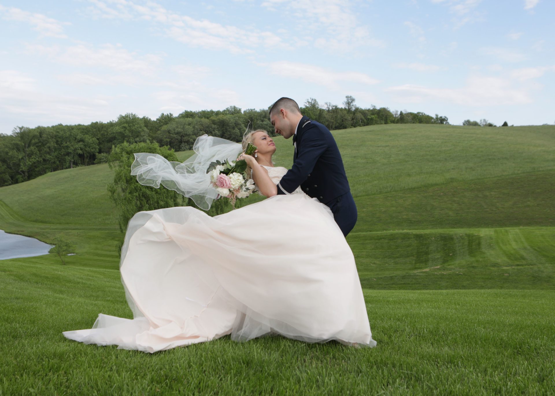 Bride and groom pose on lawn by pond at Morningside Inn with breeze lifting veil.