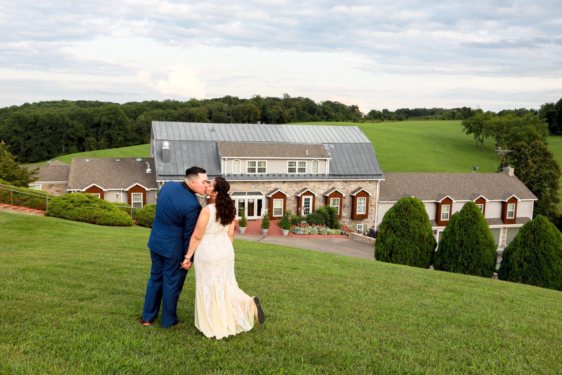 A couple kisses on a grassy hill at a picturesque wedding venue in Frederick, in front of a large building with multiple windows and a grey roof. The man wears a blue suit, and the woman wears a white dress. Trees and a cloudy sky serve as a backdrop. | Morningside Inn