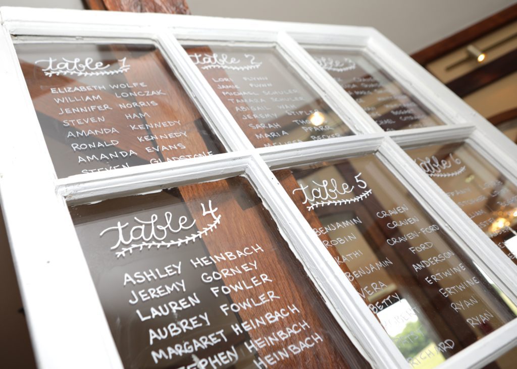 A seating chart is displayed on a vintage white window pane with multiple glass sections. Each pane lists names under different table numbers, written in white marker. Reflecting lights and wooden beams add charm to Eric & Kaitlyn's Wedding on May 28, 2017. | Morningside Inn