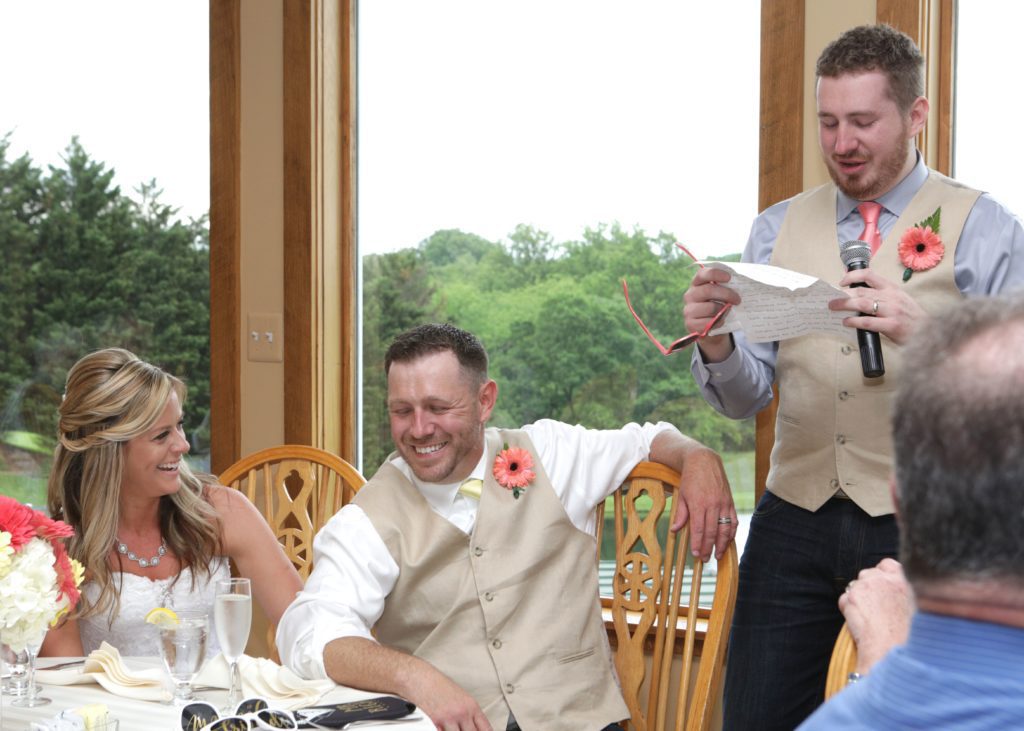 A person in a beige vest is speaking into a microphone and reading from a paper while standing next to two seated people who are laughing. They’re at Eric & Kaitlyn's Wedding, May 28, 2017, with wooden chairs, large windows, and a view of greenery outside. | Morningside Inn