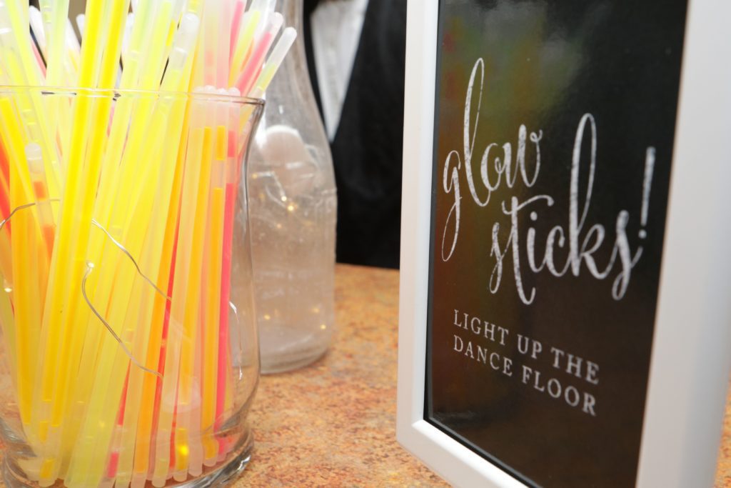 Close-up of a table with a glass vase filled with colorful glow sticks. Next to the vase is a white-framed sign with cursive writing that says, "Glow sticks! Light up the dance floor." In the background, there is a blurred figure and other objects. Celebrating Eric & Kaitlyn's Wedding, May 28, 2017. | Morningside Inn