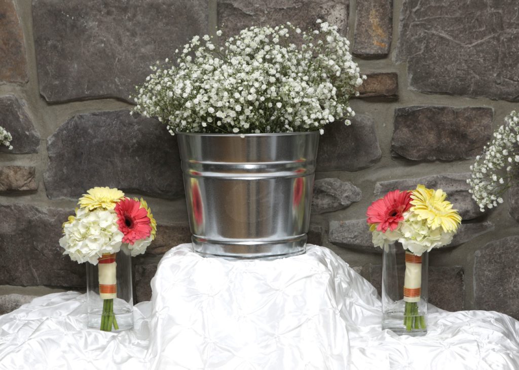 A decorative setup for Eric & Kaitlyn's wedding on May 28, featuring a metal bucket filled with white baby's breath flowers and two vases holding bouquets of pink and yellow gerbera daisies and white flowers. The display is set against a stone wall and arranged on a white, ruffled cloth. | Morningside Inn