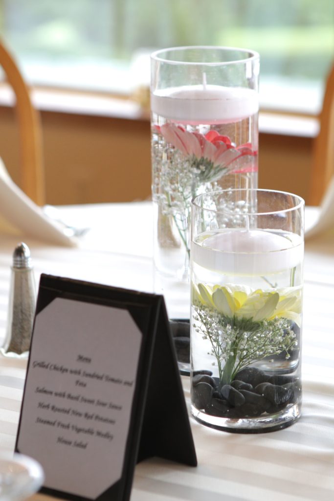 Two vases filled with water, each containing floating candles, flowers, and stones, sit on a striped tablecloth. One vase has pink and white flowers; the other yellow and white. A menu card in a black frame sits beside the vases, displaying food options for Eric & Kaitlyn's Wedding on May 28, 2017. | Morningside Inn