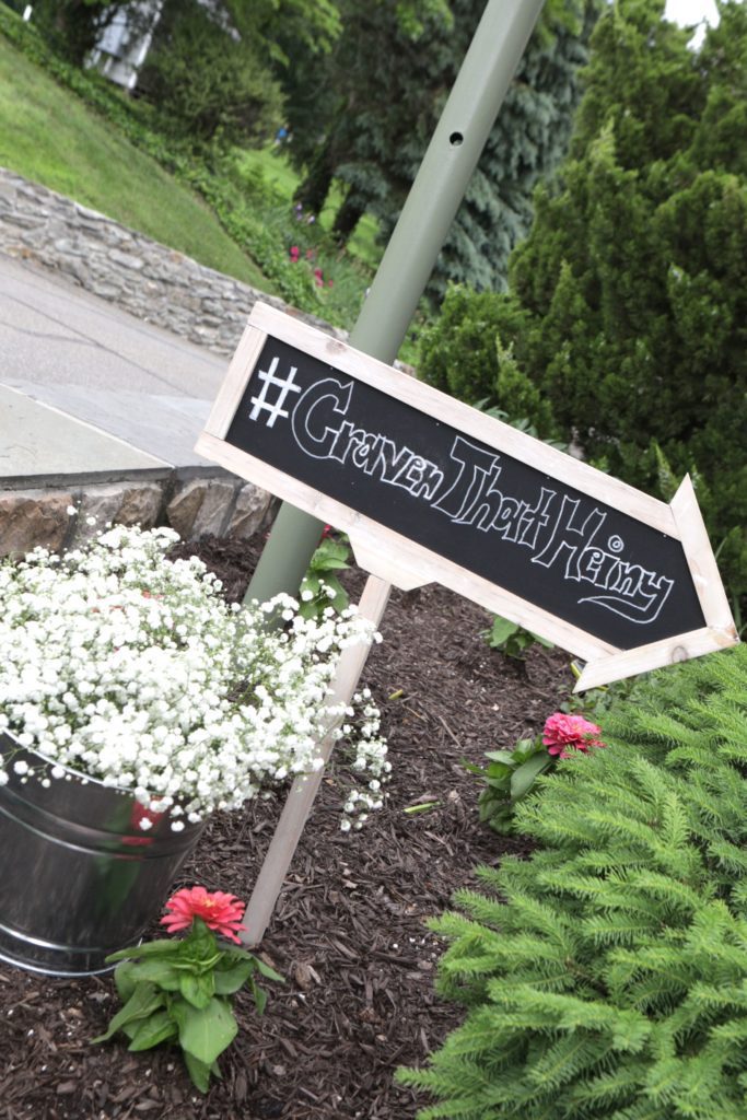 A garden scene with a chalkboard arrow sign attached to a wooden stake. The sign reads "#GravenThatHeiny" and points to the right. Below the sign is a metal bucket filled with white flowers, surrounded by green foliage and pink flowers, setting the perfect backdrop for Eric & Kaitlyn's Wedding on May 28, 2017. | Morningside Inn