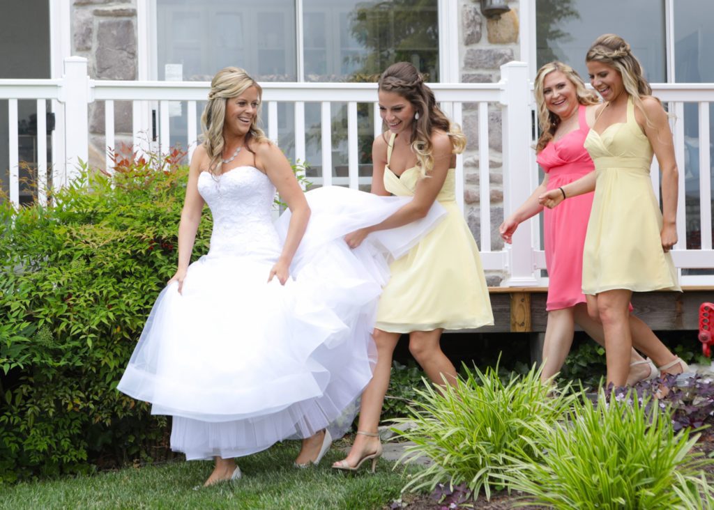 On May 28, 2017, during Eric & Kaitlyn's wedding, a bride in a white wedding dress smiles as she walks outside near a house with three bridesmaids. Two wear light yellow dresses while the other wears salmon pink. The bride holds her dress up slightly off the ground, radiating joy. | Morningside Inn