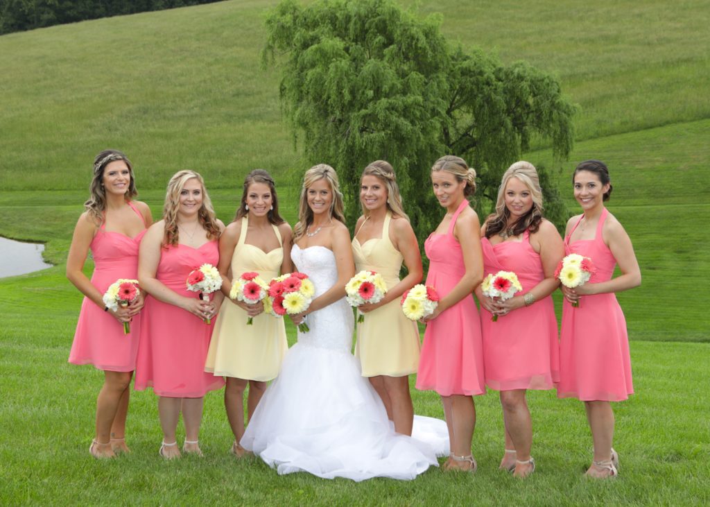 A bride in a white gown stands in the center of a grassy field, flanked by seven bridesmaids at Eric & Kaitlyn's Wedding on May 28, 2017. Four bridesmaids wear pink dresses, and three wear light yellow dresses. All hold bouquets of flowers. In the background, there is a small pond and a lush green tree. | Morningside Inn