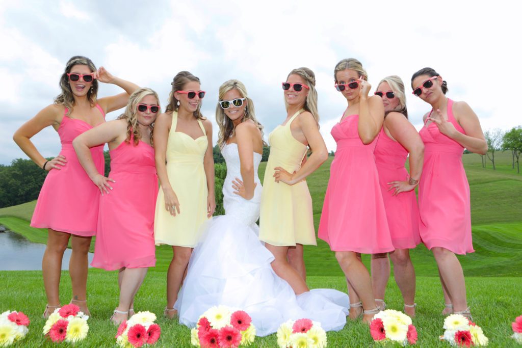 A bride in a white wedding gown is surrounded by her bridesmaids, who are wearing pink and yellow dresses. They are all wearing sunglasses and posing cheerfully on a green lawn with a scenic background of trees and a pond at Eric & Kaitlyn's Wedding on May 28, 2017. Colorful flowers are at their feet. | Morningside Inn