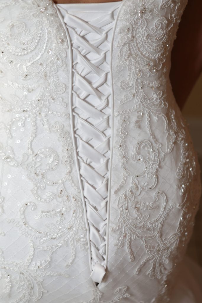 A close-up photo of a white wedding dress featuring intricate lace and bead embroidery from Eric & Kaitlyn's Wedding on May 28, 2017. The back of the dress showcases a crisscross ribbon corset design, creating a fitted and elegant look. The fabric has a subtle diagonal pattern and delicate floral details. | Morningside Inn