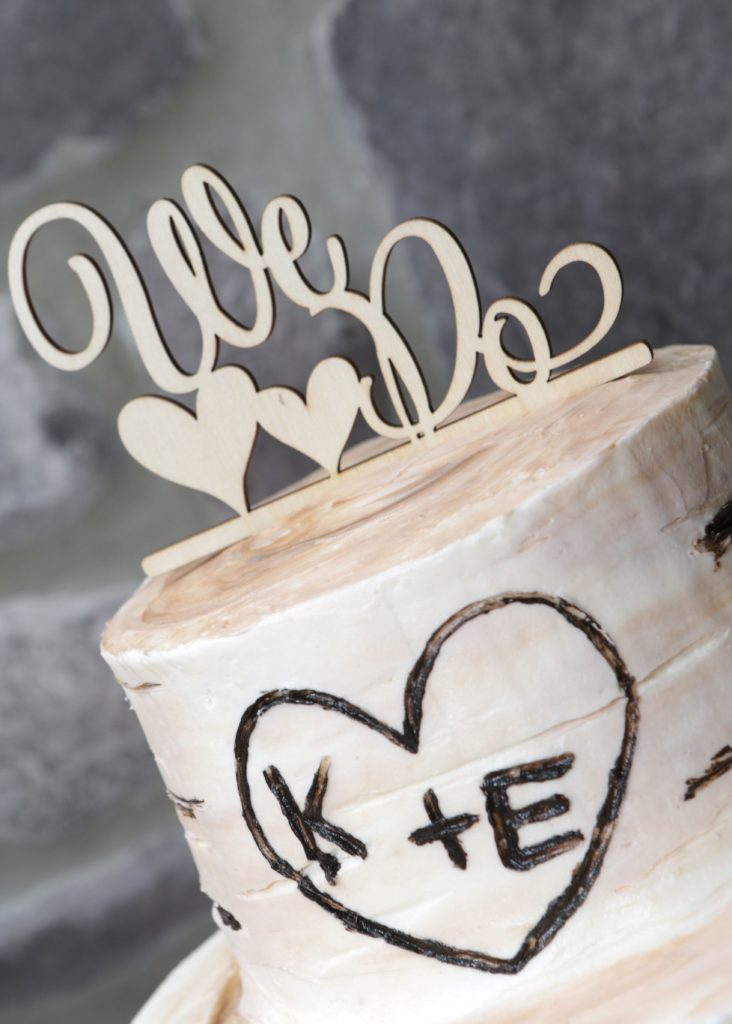 A rustic wedding cake with a carved heart featuring the initials "K+E". The top of Eric & Kaitlyn's wedding cake is adorned with a wooden topper that reads "We Do" with two hearts. The cake features a wood-like texture, enhancing its rustic theme for their special day on May 28, 2017. | Morningside Inn