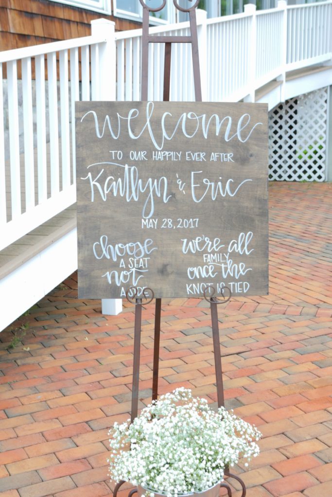 A wooden sign with white handwritten text stands on an easel on a brick path. The sign reads: "Welcome to Eric & Kaitlyn's Wedding, May 28, 2017. Choose a seat, not a side. We're all family once the knot is tied." A pot of white flowers rests beneath it. | Morningside Inn