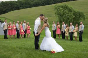 On May 28, 2017, Eric & Kaitlyn's Wedding, the bride and groom hold hands and gaze at each other, standing on a grassy hill. The bride wears a white gown and the groom a white shirt with suspenders. Their wedding party, in coral and yellow dresses and gray vests, stands behind them holding bouquets. | Morningside Inn