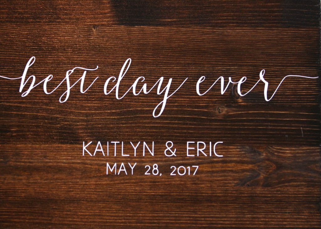 A wooden sign with elegant white calligraphy reads "best day ever" at the top. Below, it commemorates "Eric & Kaitlyn's Wedding" and "May 28, 2017" in capital letters. The background features a dark wood grain pattern. | Morningside Inn