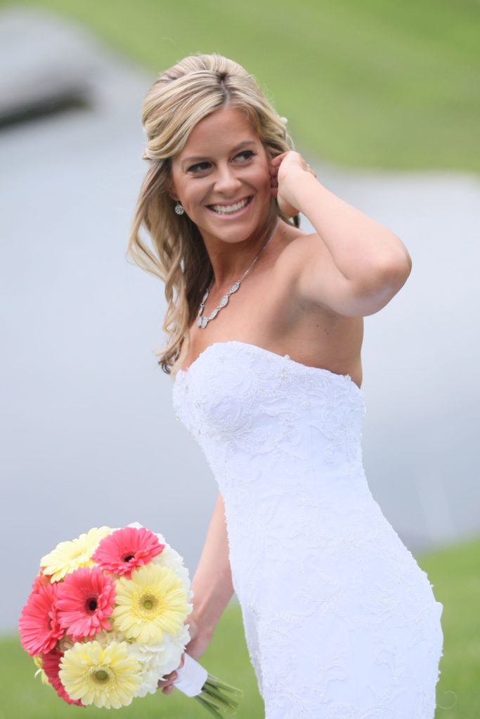 At Eric & Kaitlyn's Wedding on May 28, 2017, a bride in a strapless white dress smiles while holding a bouquet of yellow and pink flowers. Her wavy blonde hair is styled with a side part as she stands outdoors with a grassy area and water in the background. | Morningside Inn