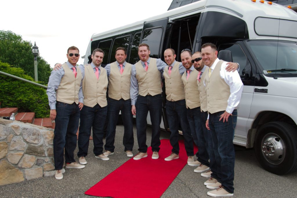 A group of eight men stand in front of a shuttle bus, dressed in matching vests, ties, shirts, jeans, and sneakers. They're lined up side by side, some with arms around each other, posing on a red carpet at Eric & Kaitlyn's wedding. The background shows greenery and a stone retaining wall. | Morningside Inn