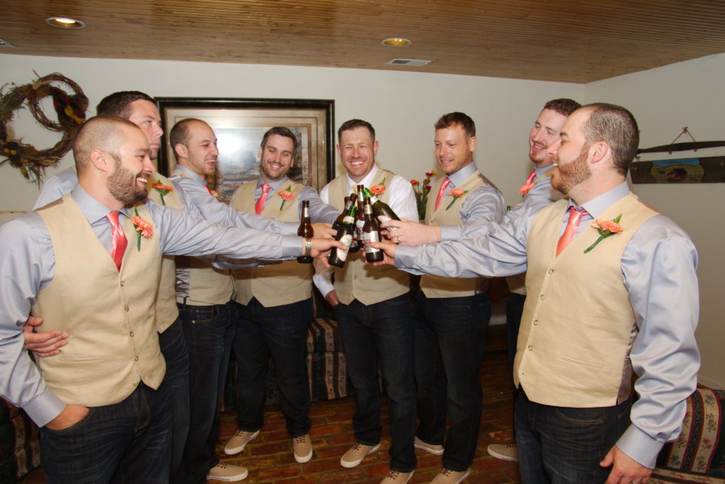 A group of men dressed in coordinated outfits, consisting of light blue shirts, beige vests, and coral ties, are clinking beer bottles together in a celebratory toast at Eric & Kaitlyn's Wedding. They are smiling and standing in a room with a wooden ceiling and framed art on the walls. | Morningside Inn