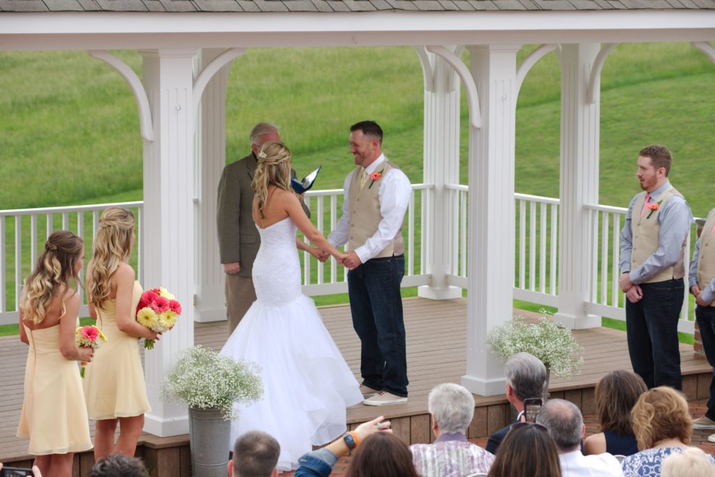 A bride and groom stand facing each other, holding hands under a gazebo during Eric & Kaitlyn's Wedding on May 28, 2017. The bride wears a white gown, and the groom wears a white shirt with a beige vest. Bridesmaids in light yellow dresses and groomsmen in light vests stand nearby. | Morningside Inn