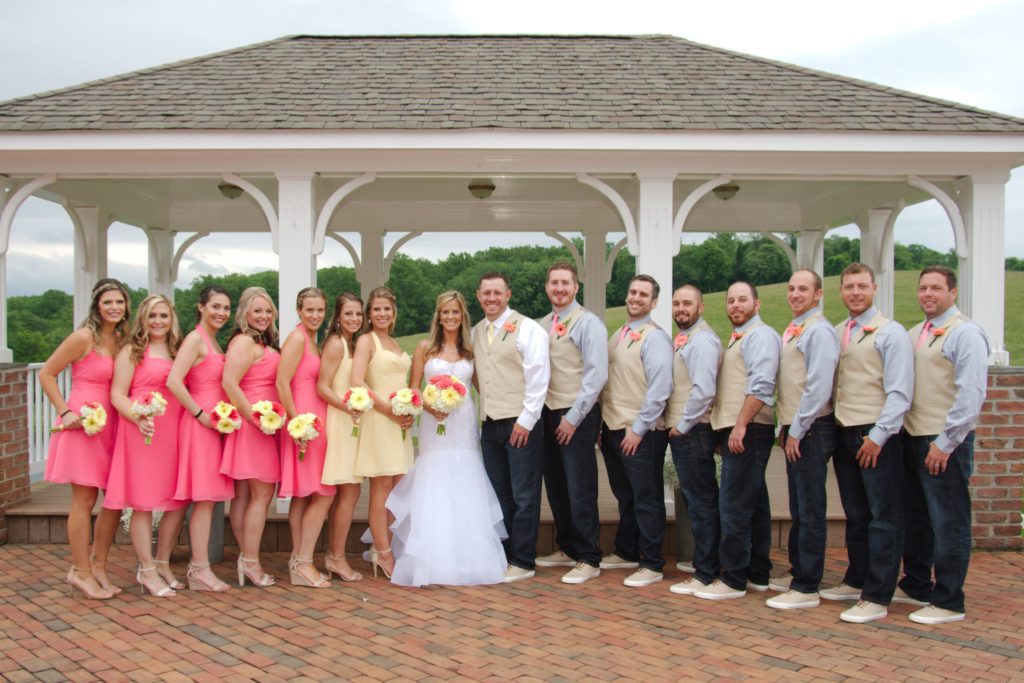 On May 28, 2017, Eric and Kaitlyn's wedding party poses in front of a gazebo. The bride and groom stand at the center, with bridesmaids in pink dresses holding yellow bouquets on the left and groomsmen in gray vests with peach boutonnieres on the right. Greenery decorates the background. | Morningside Inn