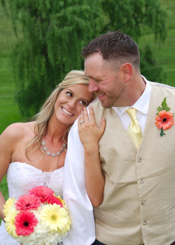 A smiling bride in a strapless white gown and groom in a beige vest with a yellow tie stand outdoors. She holds a bouquet of pink, white, and yellow flowers. They lean close to each other lovingly, surrounded by lush greenery on Eric & Kaitlyn's Wedding, May 28, 2017. | Morningside Inn