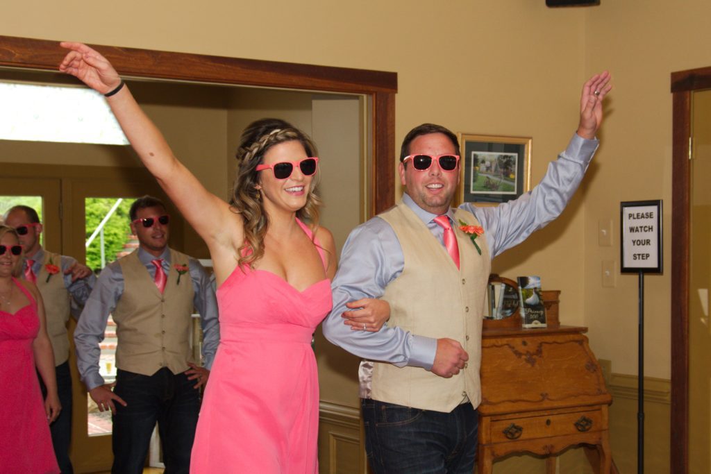 A cheerful woman and man, both wearing sunglasses and pink attire, make their entrance into a room with large smiles and raised arms. The woman wears a pink dress, and the man sports a pink tie, light vest, and jeans. A "Please Watch Your Step" sign is seen in the background at Eric & Kaitlyn's Wedding on May 28, 2017. | Morningside Inn