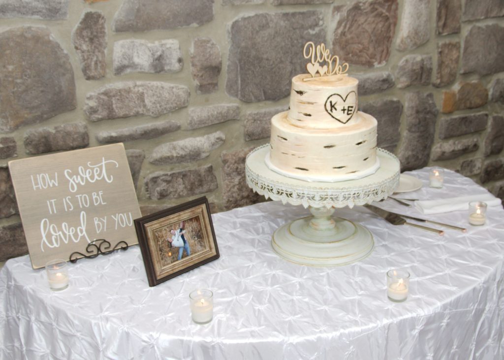 A rustic two-tier wedding cake with a wood texture design on a white pedestal stand. The cake topper reads "We Do" with initials "K + E" carved into a heart. On the table are candles, a framed photo of the couple, and a sign reading "How sweet it is to be loved by you." Eric & Kaitlyn's Wedding, May 28, 2017. | Morningside Inn