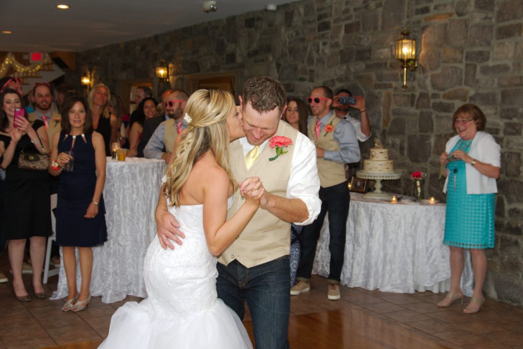 At Eric & Kaitlyn's Wedding on May 28, 2017, a couple shares a dance at their reception. The bride, in a white gown, and the groom, in a beige vest and jeans, embrace closely. Guests in casual attire watch and some take pictures with a stone wall and cake table in the background. Everyone appears joyous. | Morningside Inn