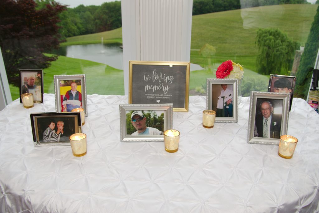 A memorial table draped in white fabric displays framed photos of deceased loved ones, with lit candles placed among them. The backdrop is a scenic view of a grassy field and pond. A central sign reads "In loving memory" with names of the individuals honored at Eric & Kaitlyn's Wedding on May 28, 2017. | Morningside Inn