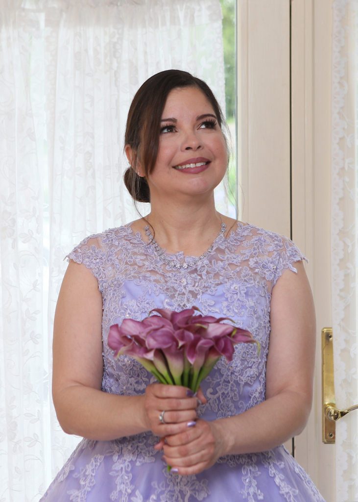 At Maria & Mike's Wedding on May 6, 2017, a woman with dark hair in an updo, wearing a lilac lace dress, smiles while holding a bouquet of purple calla lilies. She stands indoors in front of a glass door with white lace curtains, as light from outside illuminates the scene. | Morningside Inn