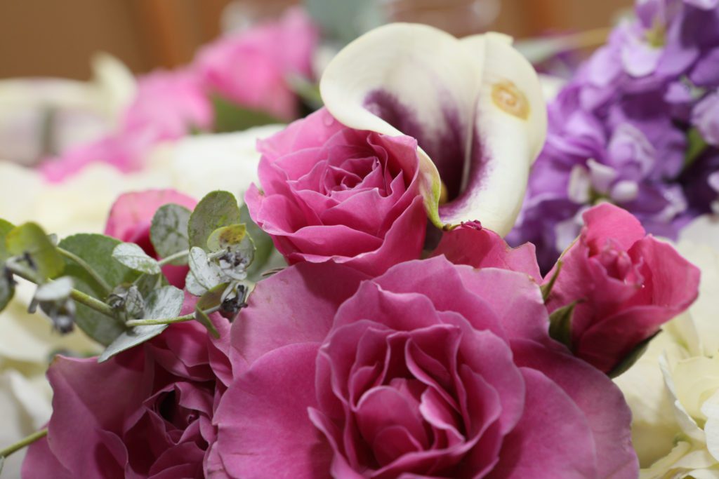 A close-up of a floral arrangement from Maria & Mike's Wedding on May 6, 2017, featuring vibrant pink roses, white calla lilies with purple centers, and sprigs of eucalyptus leaves. The background shows blurred purple and white flowers, adding depth to the bouquet. | Morningside Inn