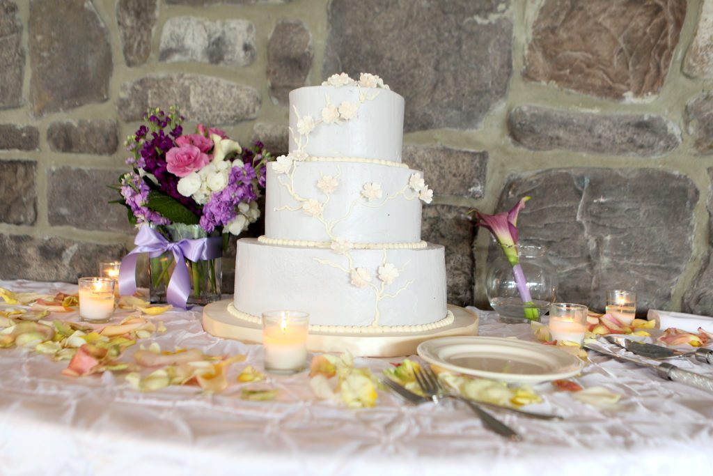 A three-tier white wedding cake with floral decorations, placed on a table with scattered rose petals and lit candles. To the left of the cake is a bouquet of pink, white, and purple flowers in a vase. The background features a stone wall, setting the perfect scene for Maria & Mike's Wedding on May 6, 2017. | Morningside Inn