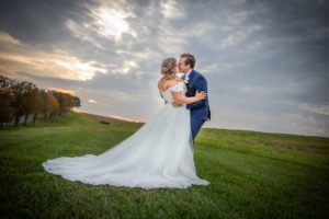 A bride and groom embrace on a grassy hillside with trees and a sunset sky behind them.