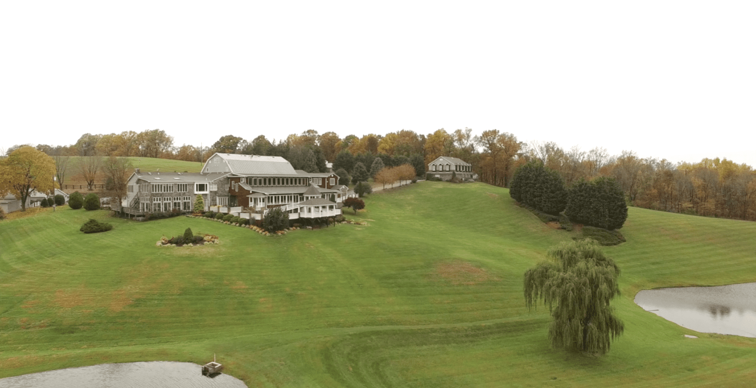 Aerial view of a rural landscape featuring a large house with multiple annexes and porches, surrounded by a wide expanse of lawns, a few rows of autumn trees, and a small pond.