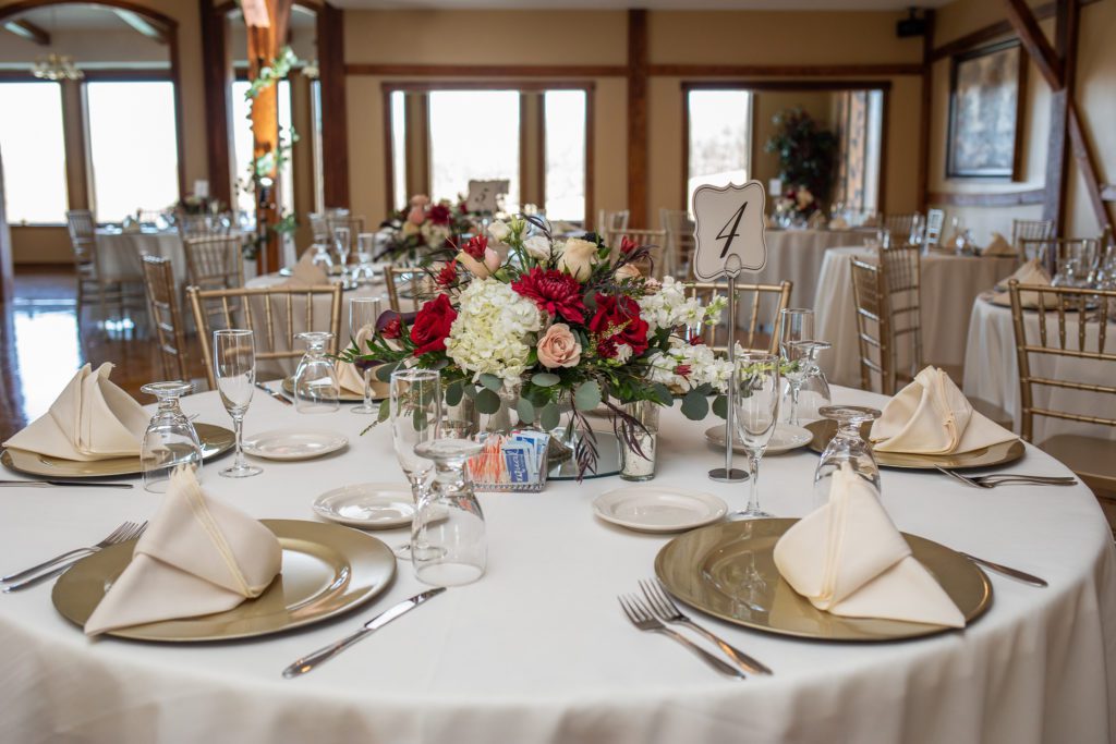 Elegant wedding reception table setting with gold chargers, beige linens, fine silverware, white and red floral centerpiece, and table number sign in a banquet hall.