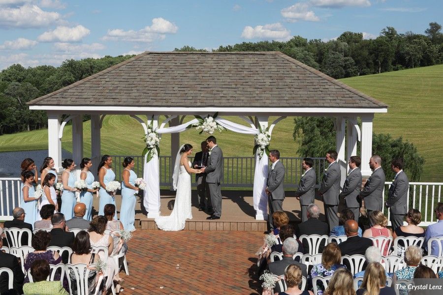 An outdoor wedding ceremony taking place under a white gazebo, with a bride and groom standing at the altar facing each other and a wedding officiant nearby. To the sides are bridesmaids in blue dresses and groomsmen in grey suits, with seated guests in the foreground all facing the gazebo. The setting is on a well-manicured lawn with a body of water and trees in the background.