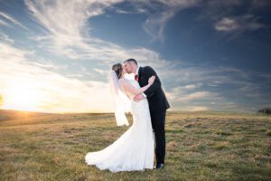 A bride and groom kissing in an open field with a vivid sky and sunset in the background.