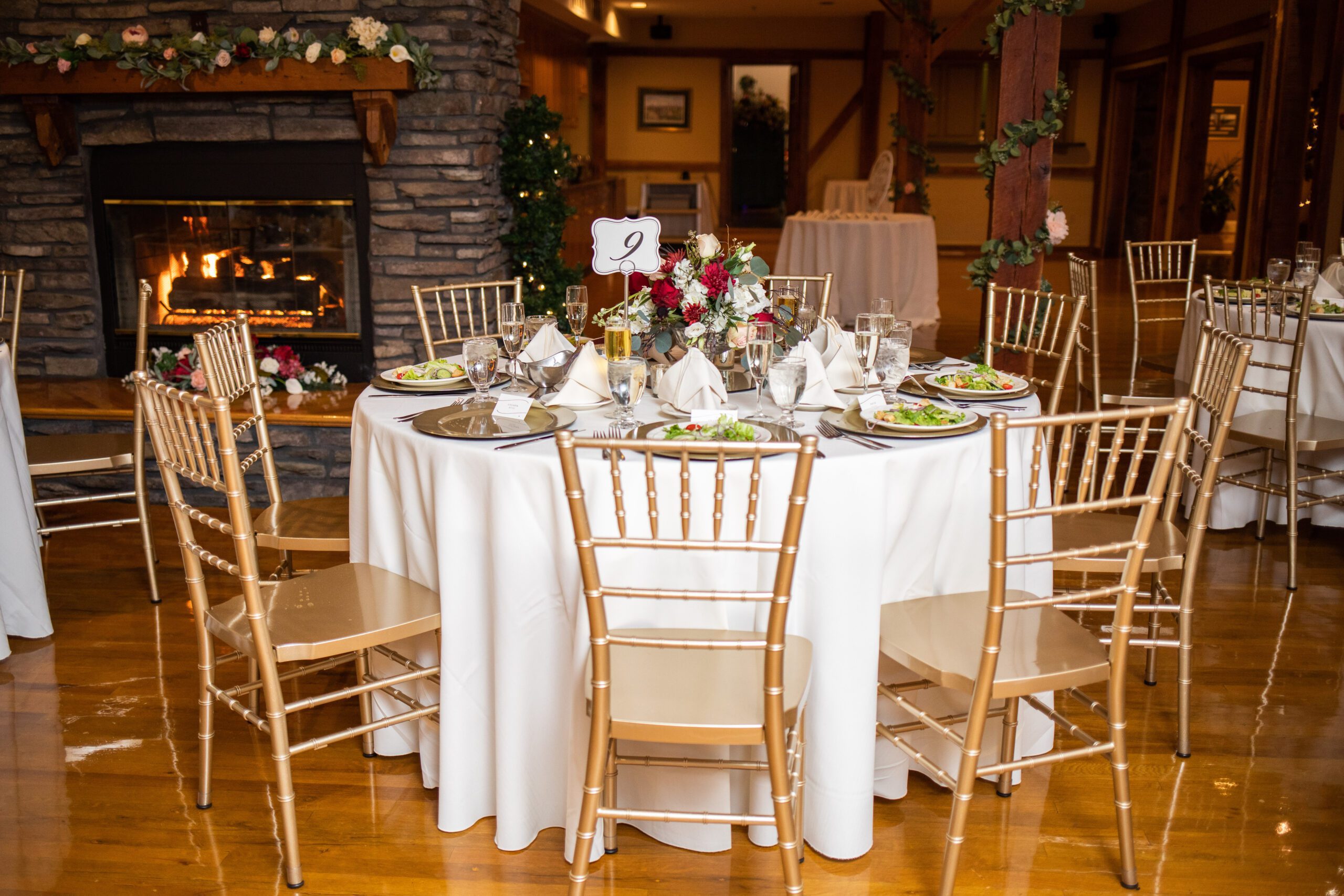 An elegantly set dining table at a formal event with gold Chiavari chairs, white tablecloths, dinner plates, and a floral centerpiece, with a lit fireplace in the background.