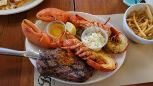 A surf and turf meal featuring a grilled steak, lobster tail, whole cooked crab, shrimp, a side of coleslaw, melted butter with a lemon wedge, and a separate bowl of french fries on the table.