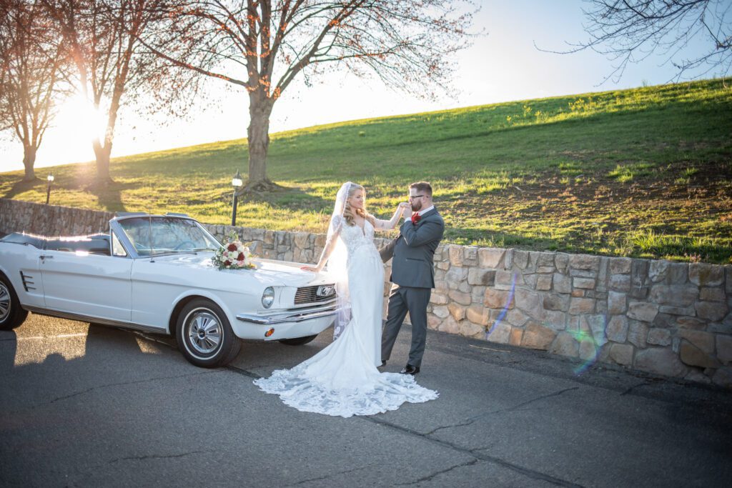 A bride and groom sharing a moment beside a white vintage convertible car decorated with flowers, with a backdrop of a sunlit park setting.