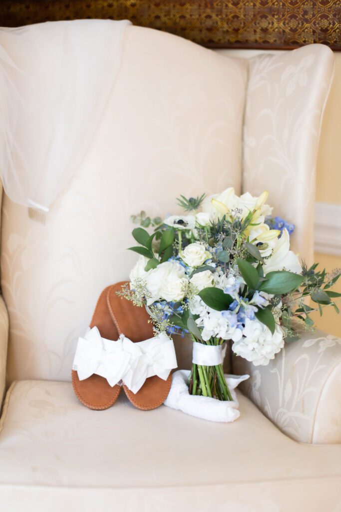 A bridal bouquet with white roses, blue flowers, and greenery, paired with white bridal sandals with large bows, resting on an elegant cream-colored armchair.